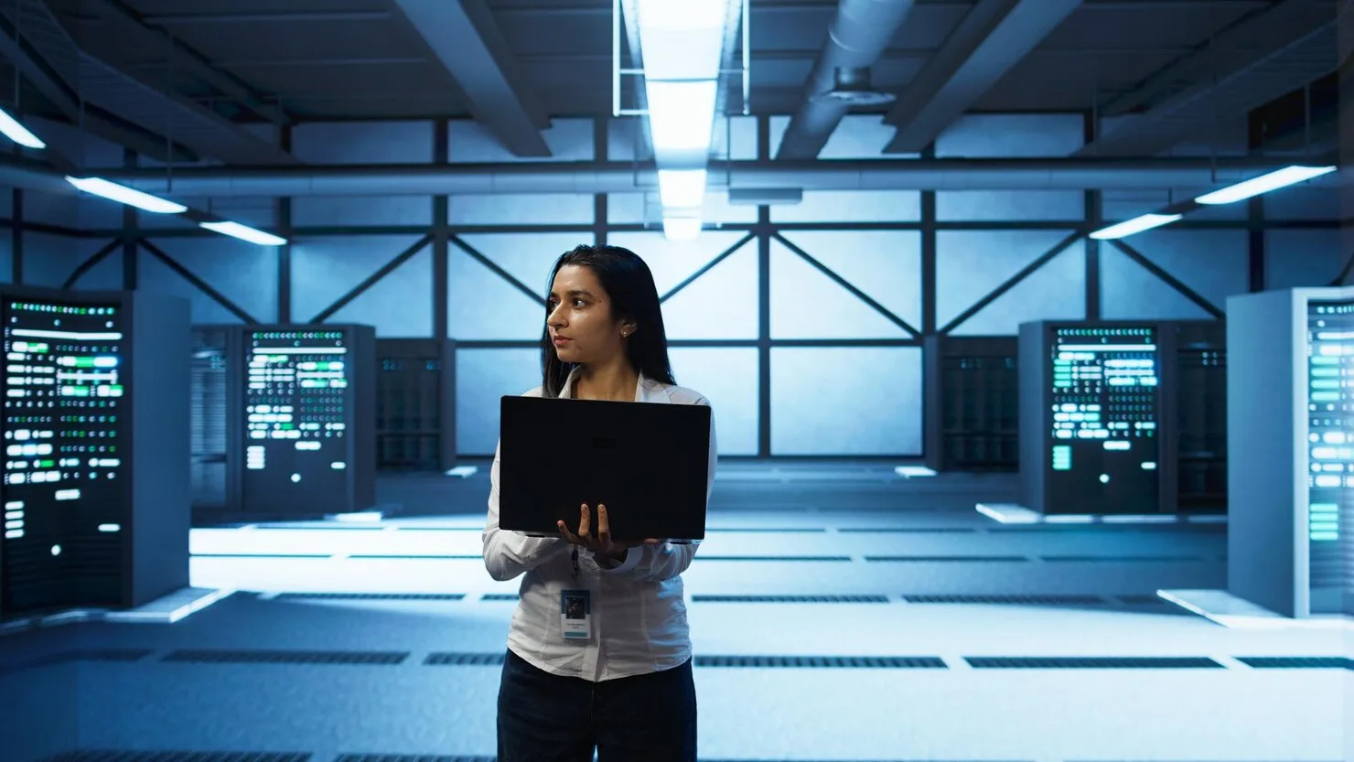 A unosquare technical expert standing in a modern data center holding a laptop, surrounded by illuminated server racks.